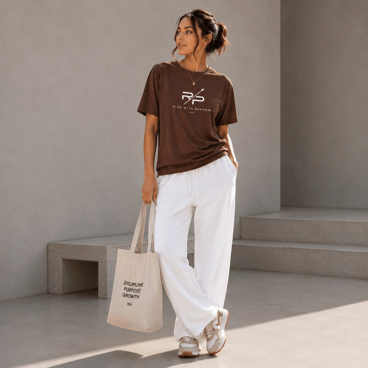 Rise With Purpose brown Sorona t-shirt with logo, paired with white wide-leg pants and sneakers. Model holds a canvas tote bag.